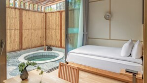 A modern hotel room with a wooden floor, a bed with white linens, a small wooden table with a vase and plant, and a hot tub in the background.