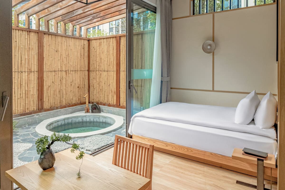 A modern hotel room with a wooden floor, a bed with white linens, a small wooden table with a vase and plant, and a hot tub in the background.