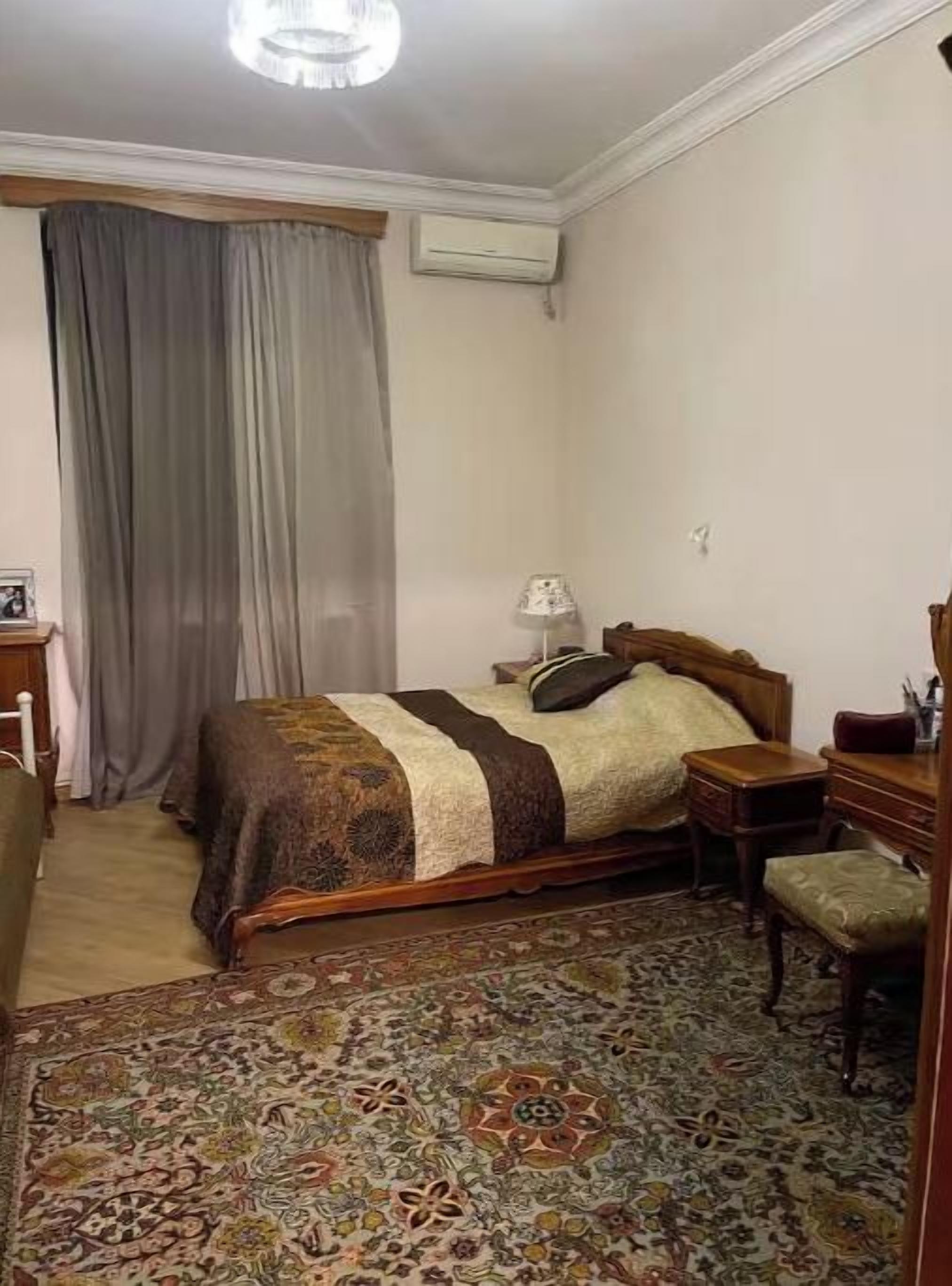Un dormitorio con cama, mesita de noche, silla, escritorio y una alfombra estampada.