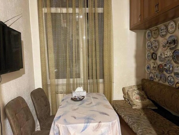 A room with a dining table covered by a tablecloth, two chairs, a sofa, a television mounted on the wall, and a curtain.