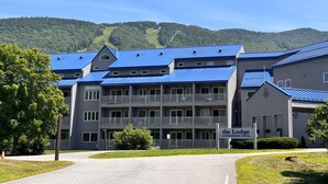 A multi-story building with a blue roof, a sign reading "The Lodge Conference Hotel," and a backdrop of green hills. - 1BR | Balcony over the Pemi River | Loon Views | Indoor Pool & Hot Tub (Lincoln)