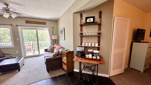 A cozy living room with a sofa, a coffee table, a dining table with a coffee maker, and a refrigerator. There is a sliding glass door leading to a balcony. - 1BR | Balcony over the Pemi River | Loon Views | Indoor Pool & Hot Tub (Lincoln)
