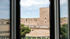 A view of a historic stone building with multiple towers and arched windows, viewed through a window with a metal railing. - Palazzina Gentile (Bari)