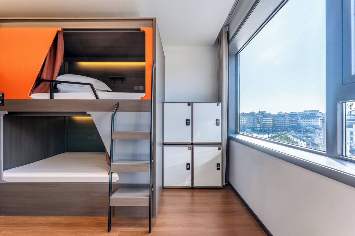 Une chambre d’hôtel moderne équipée d’un lit superposé, d’une petite kitchenette et d’une grande fenêtre donnant sur la ville.