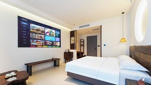 Una habitación de hotel moderna con una pantalla digital grande en la pared, una cama bien hecha, un cabecero de madera, una mesita de noche y un banco.