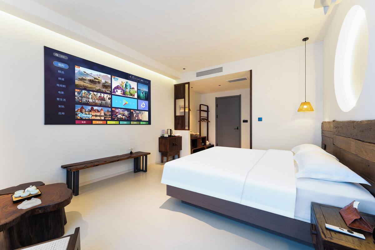 Une chambre d’hôtel moderne avec un grand écran numérique au mur, un lit parfaitement fait, une tête de lit en bois, une table de chevet et un banc.