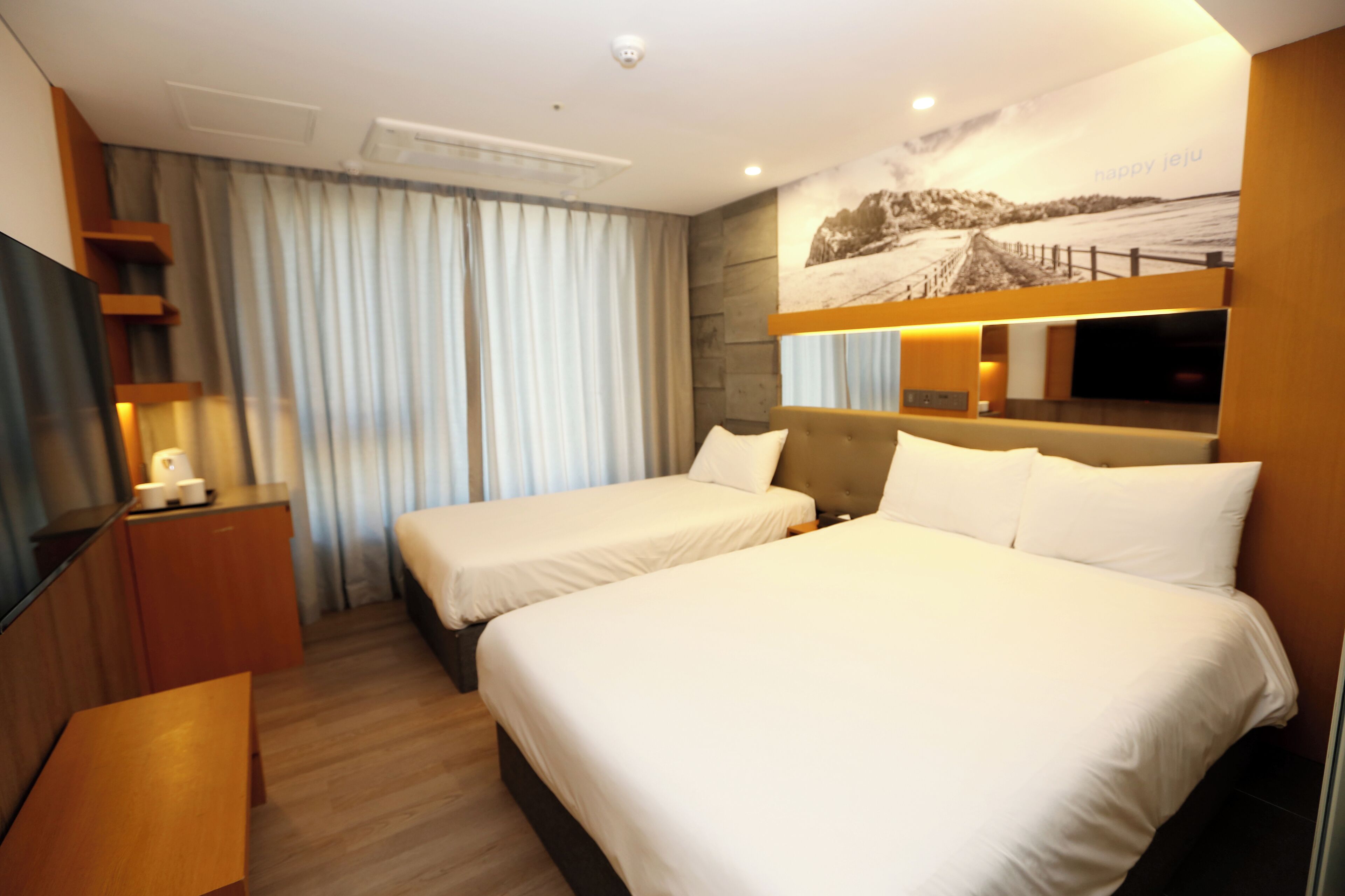 A hotel room with two beds, a desk, a TV, and a large window with curtains.