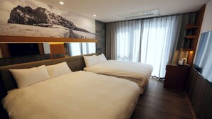 Ein Hotelzimmer mit zwei Betten, einem großen, an der Wand befestigten Fernseher, einem Berglandschaftsbild und einem Fenster mit Vorhängen.