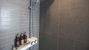 Ein Badezimmer mit dunkel gefliesten Wänden, einer Dusche mit Handbrause und drei Shampoo-Flaschen auf einem Regal.