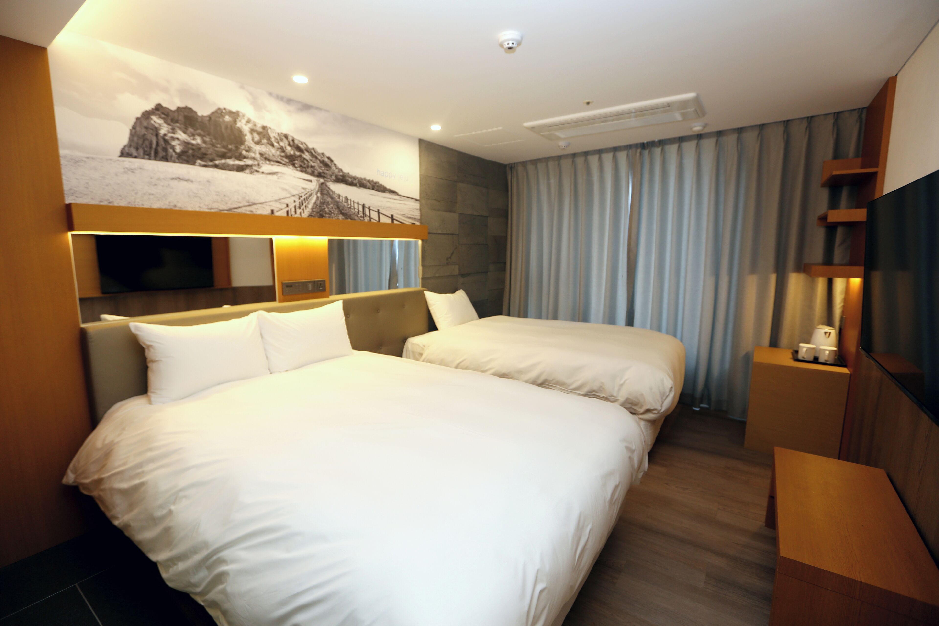 A hotel room with two beds, a large wall-mounted TV, a flat-screen TV, a wooden headboard, a bedside table, and a window with curtains.