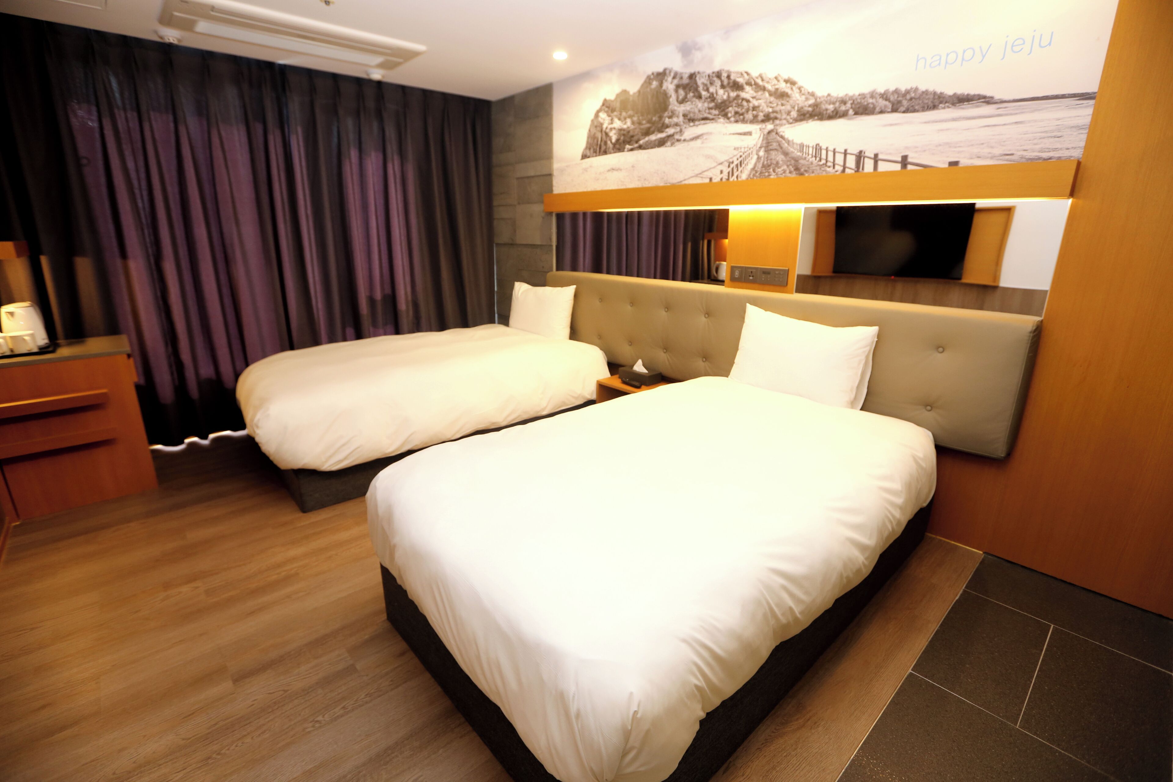 A hotel room with two beds, a wooden desk, a flat-screen TV, and a large window with curtains.
