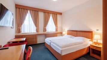 Habitación de hotel con cama doble, escritorio con silla, televisor y ventana con cortinas.