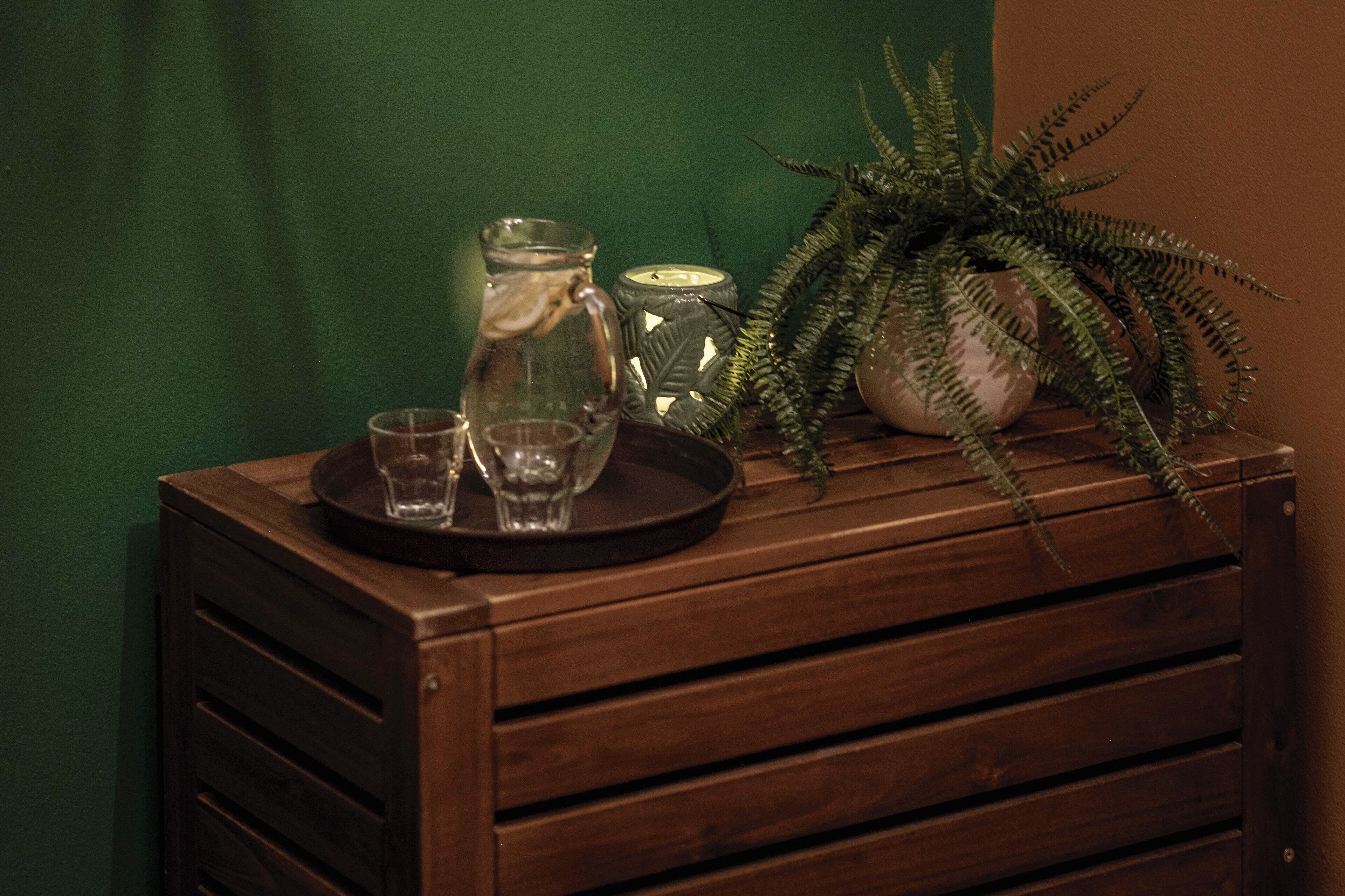 Un mueble de madera con una jarra de cristal, una vela y una planta de helecho encima.