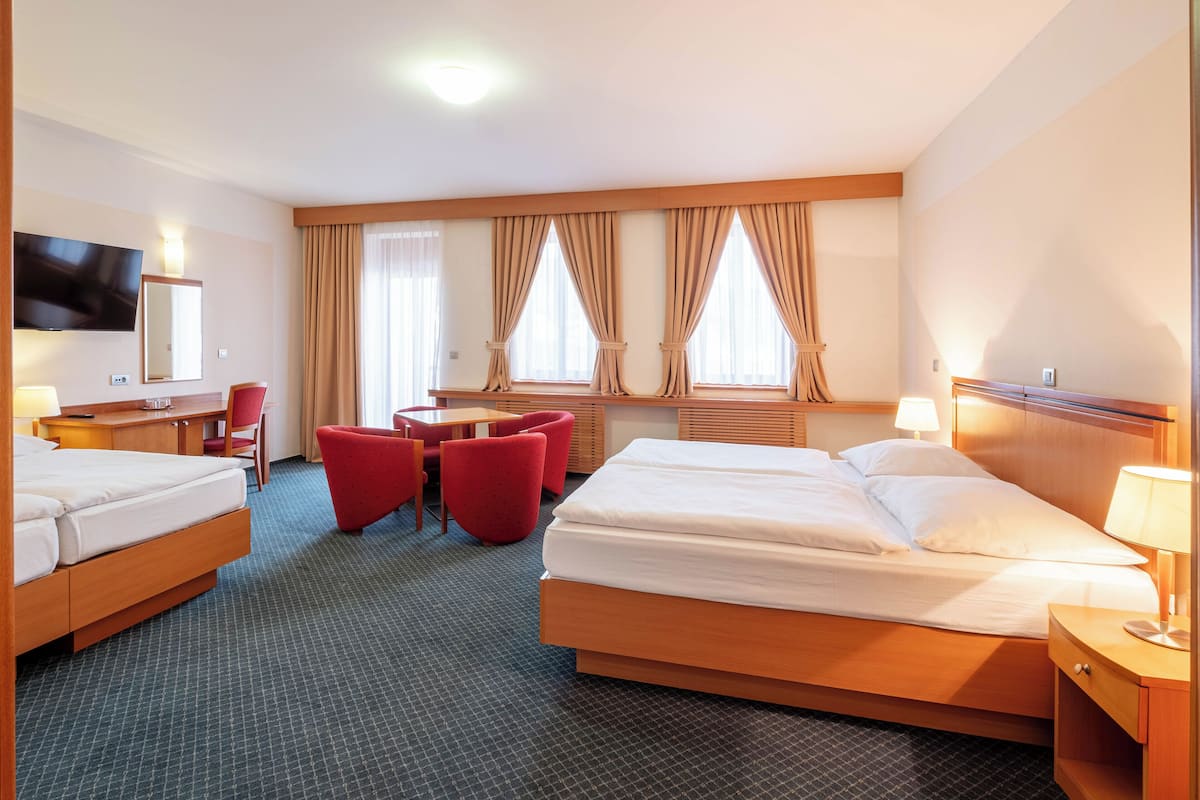 Habitación de hotel con dos camas, un escritorio, una silla, un televisor y ventanas amplias con cortinas.