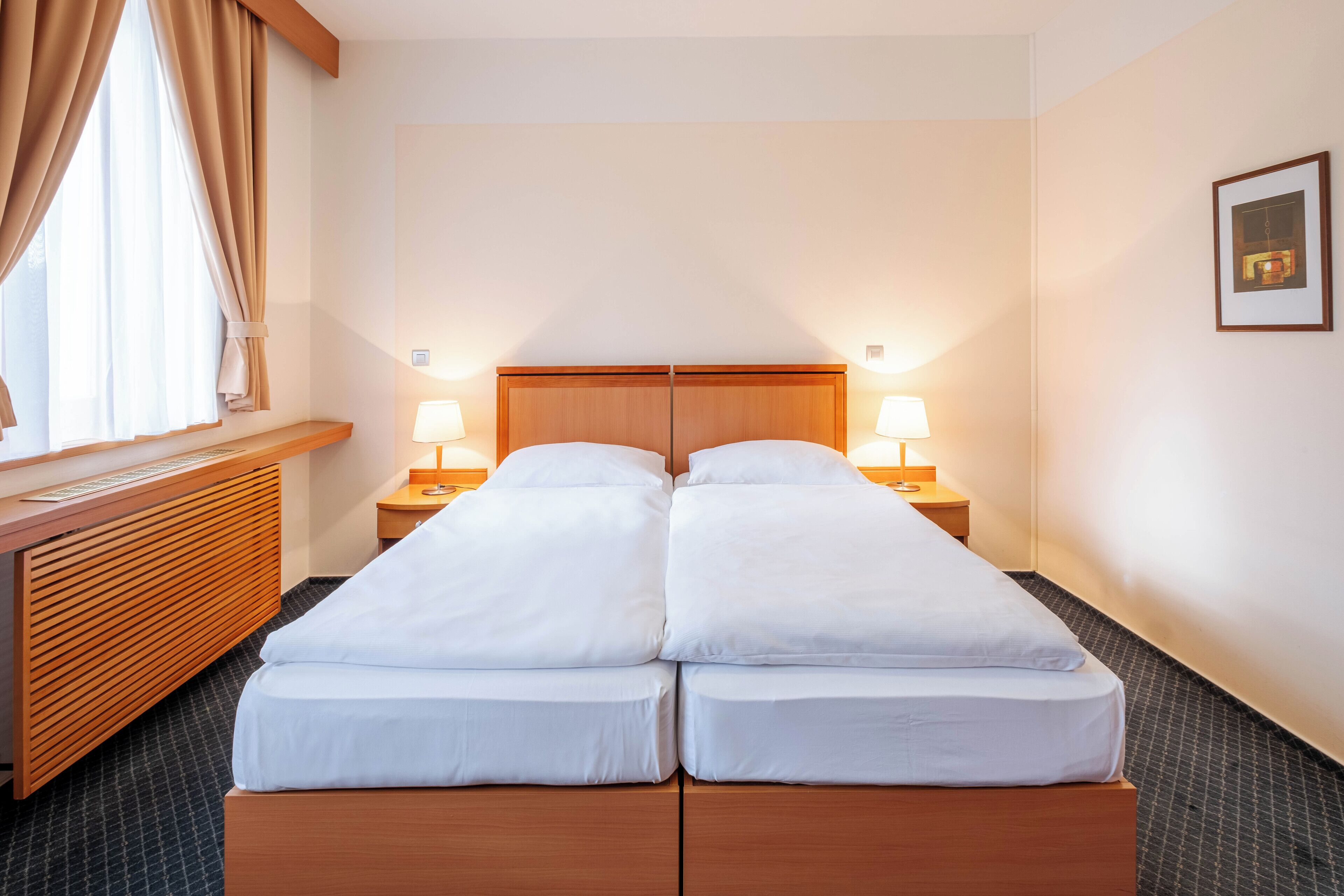 A double bed with wooden headboard, two bedside tables with lamps, a window with curtains, and a framed picture on the wall.