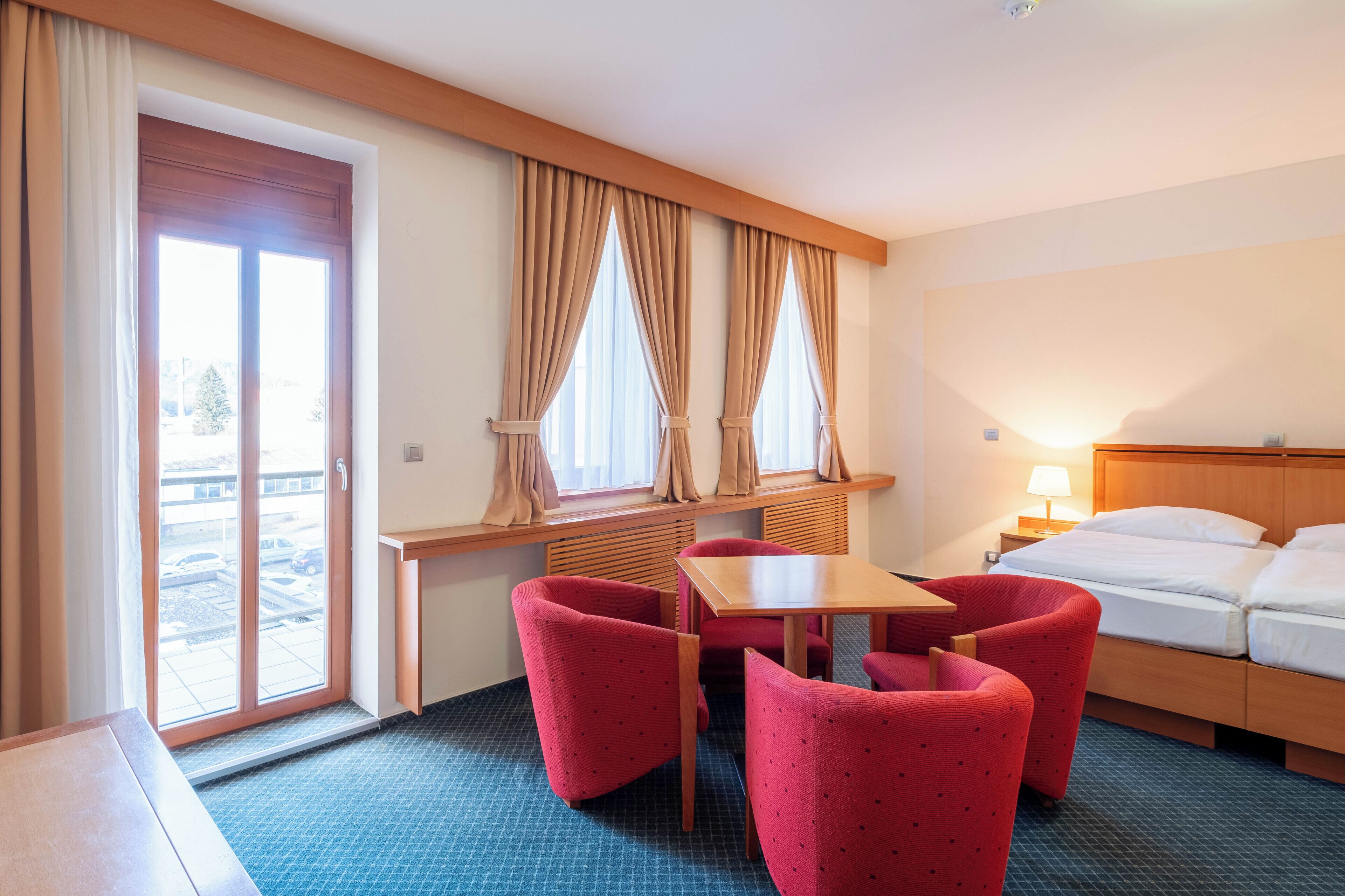 Ein Hotelzimmer mit einem Bett, einem Schreibtisch, zwei roten Stühlen, einem Fenster mit Vorhängen und Blick auf einen Parkplatz.