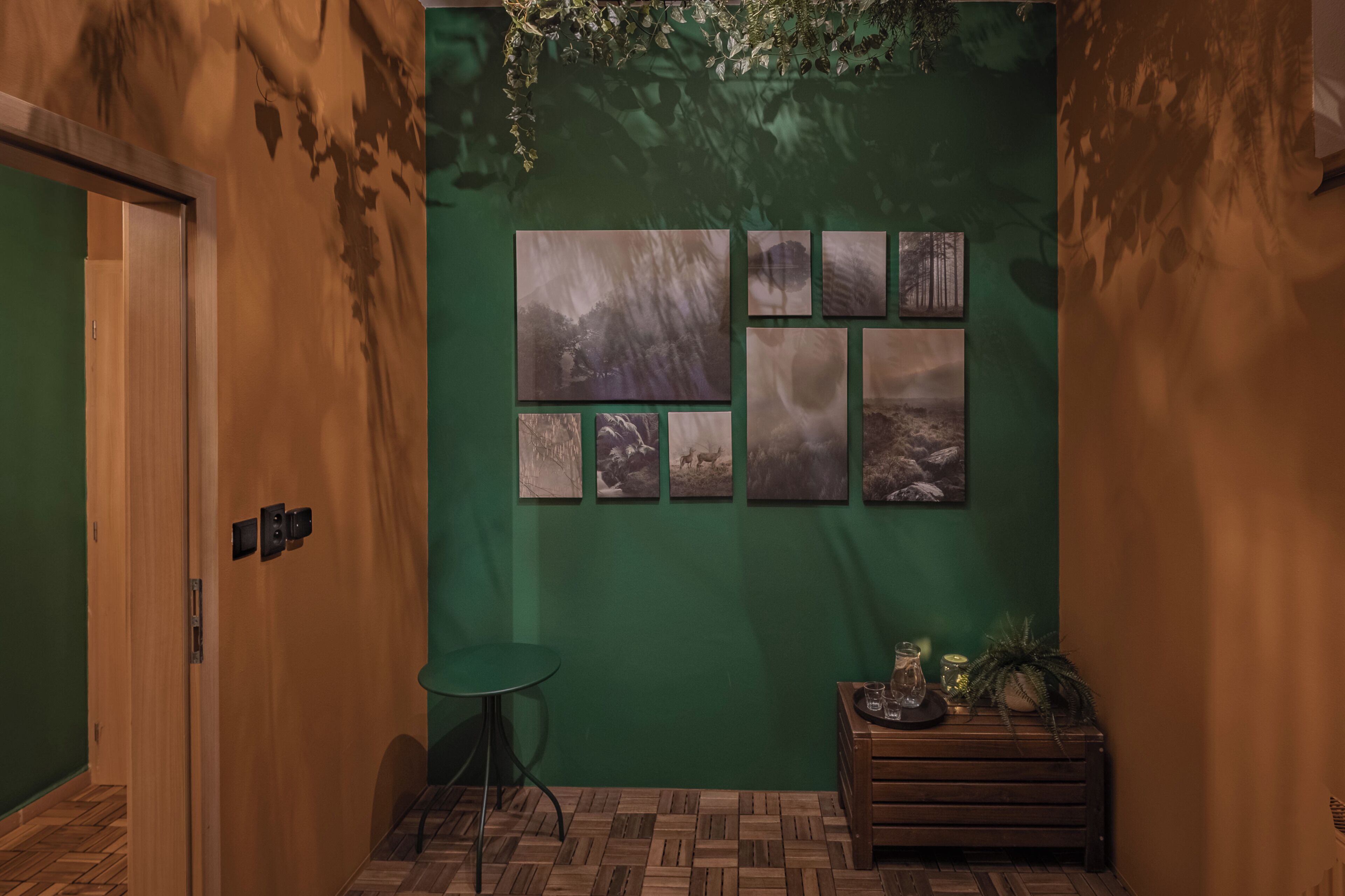 A hallway with a green wall, wooden floor, and a series of framed pictures. There is a door on the left, a small round table, and a wooden bench with a plant on it.