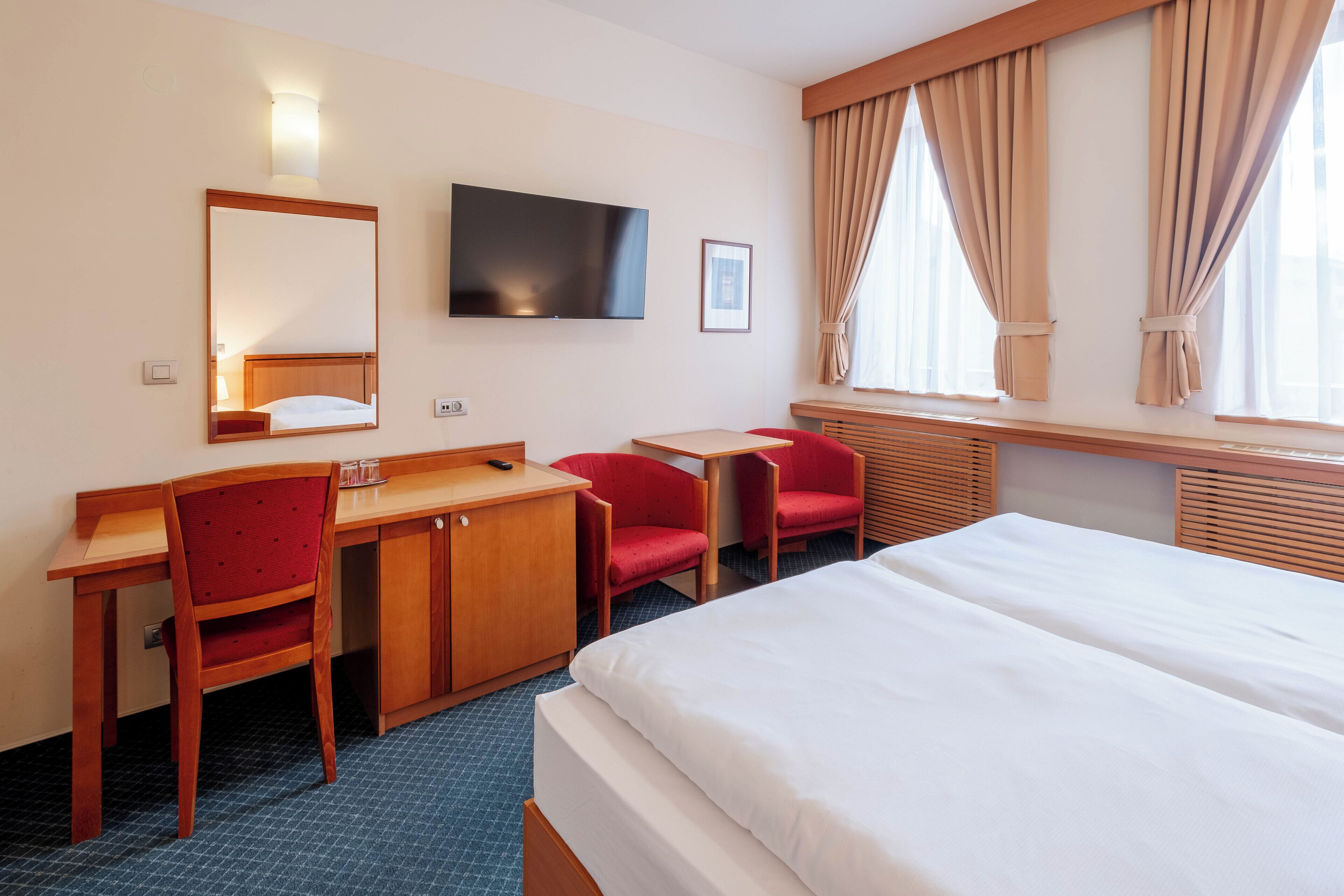 A hotel room with a bed, a desk, two red chairs, a TV, and a window with curtains.