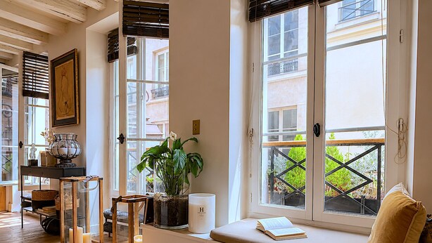 A room with a large window, a bench with a cushion, a potted plant, a candle, and a small table with a lamp. - L'appartement Charlot L (Paris)