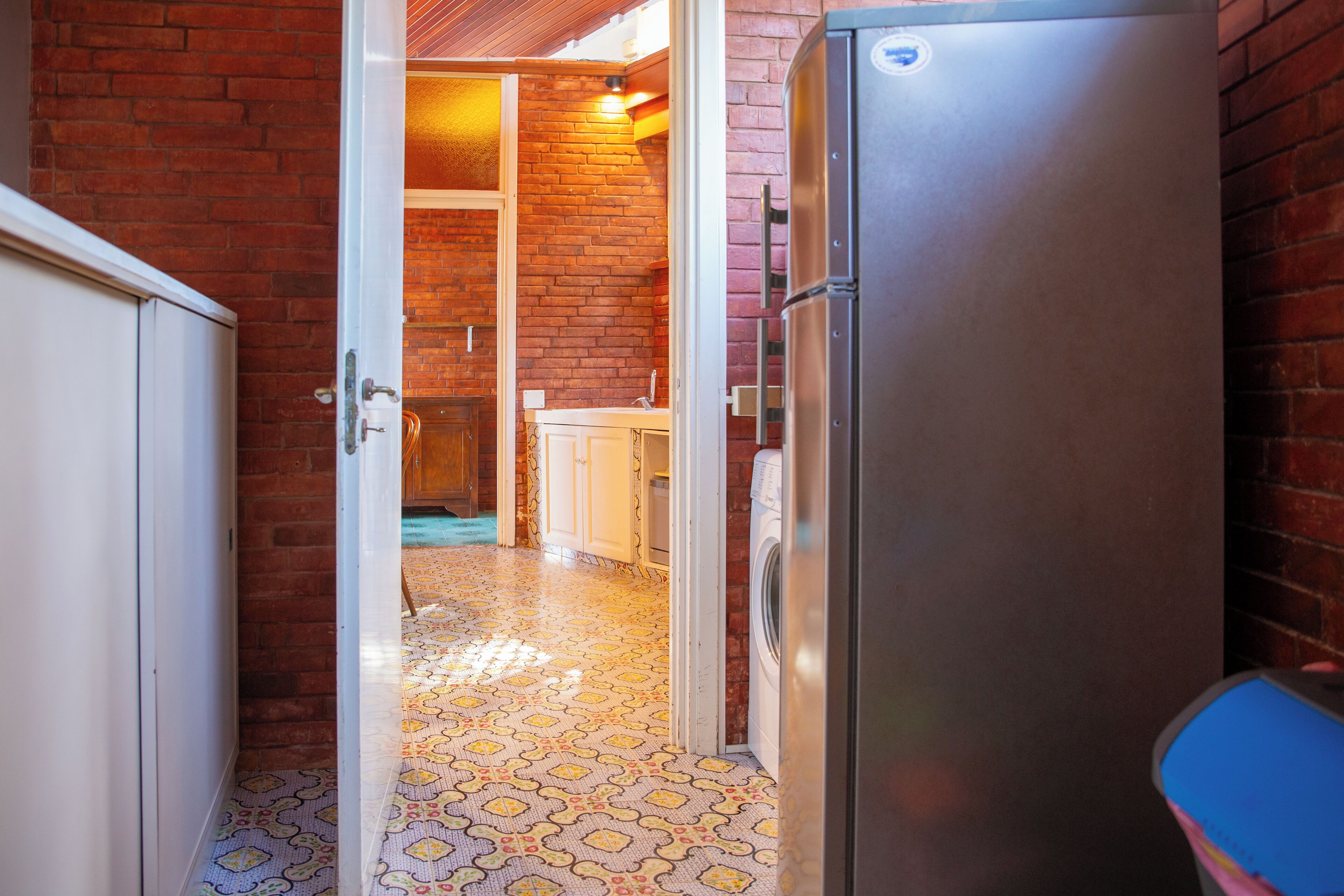 Un couloir étroit, dont le sol est recouvert d’un motif décoratif, mène à une cuisine équipée d’un réfrigérateur et d’un lave-linge.