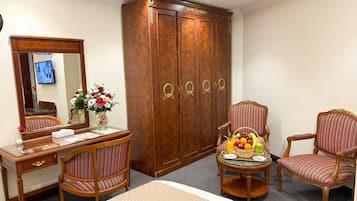 Une chambre d'hôtel avec une armoire en bois, un bureau avec un miroir, deux chaises à rayures et une petite table sur laquelle se trouvent un panier de fruits et des boissons.