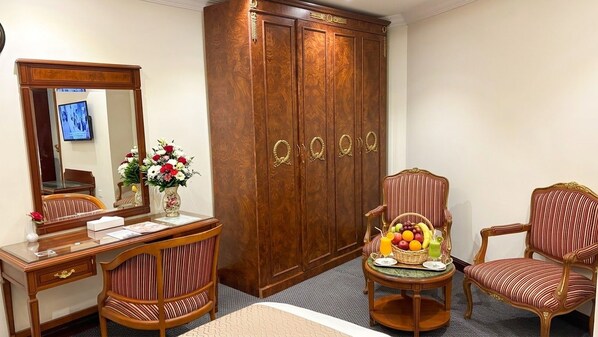 A hotel room with a wooden wardrobe, a desk with a mirror, two striped chairs, and a small table with a fruit basket and drinks. - Taj Celine Hotel (Jeddah)