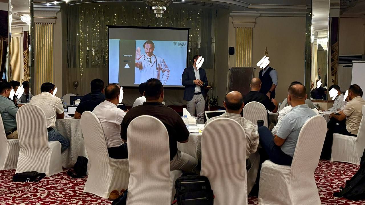 A conference room with attendees seated at round tables, a presentation screen, and a speaker at the front.