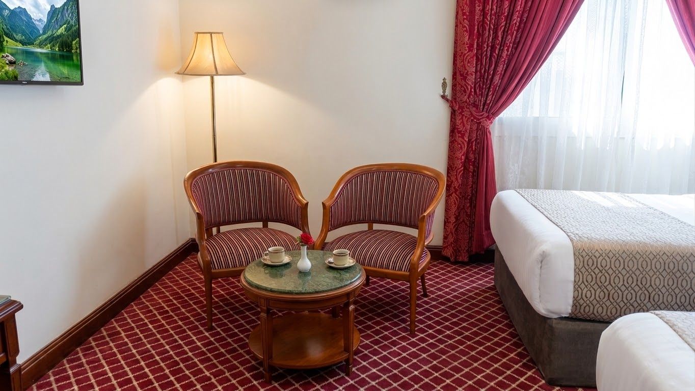 A hotel room with a bed, two upholstered chairs, a small round table with a vase and two cups, a lamp, and a painting on the wall.
