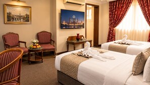 Een hotelkamer met twee bedden, een eettafel met fruit, twee gestreepte fauteuils, een schilderij van een stadsgezicht en een raam met gordijnen.