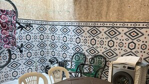A dining area with a table covered by a dark cloth, surrounded by white plastic chairs. A washing machine is visible in the background. - Apartment sidi rahhal near beach (Sidi Rahal)