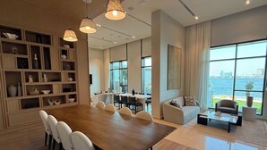 A spacious living area with a large wooden dining table, white chairs, a sofa, and a coffee table. There are shelves with decorative items and a view of the water through large windows.