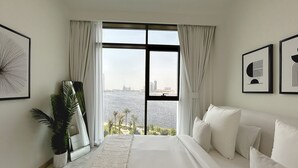 A bedroom with a large window, white bedding, a potted plant, and framed artwork on the walls.