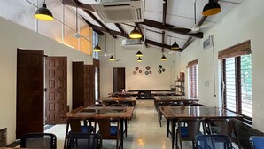 Une salle de classe avec des tables et des chaises en bois, des ventilateurs de plafond et des lampes suspendues.