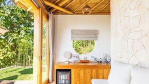 Une chambre avec un bureau en bois, un petit réfrigérateur, un canapé blanc et une vue sur un jardin.
