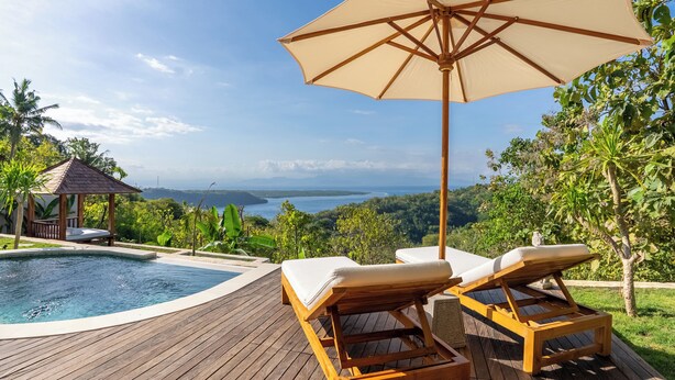 Une terrasse en bois avec deux chaises longues et un grand parasol, offrant une vue sur une piscine et un paysage pittoresque.