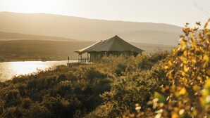 Une structure en forme de tente, dotée d’un balcon, entourée de végétation naturelle et bordée d’un plan d’eau, avec des collines en arrière-plan.