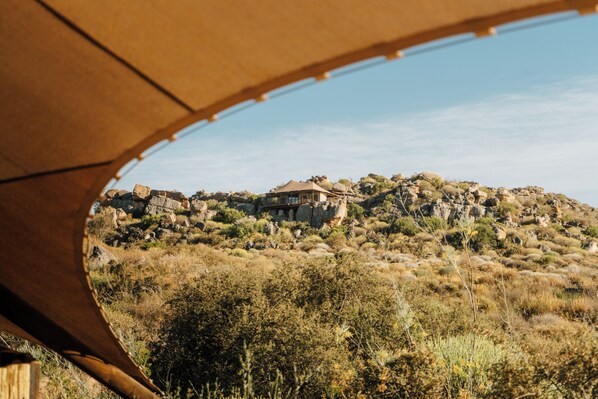 Une vue à travers une structure en bois, avec une maison sur une colline, entourée d’une végétation aride.