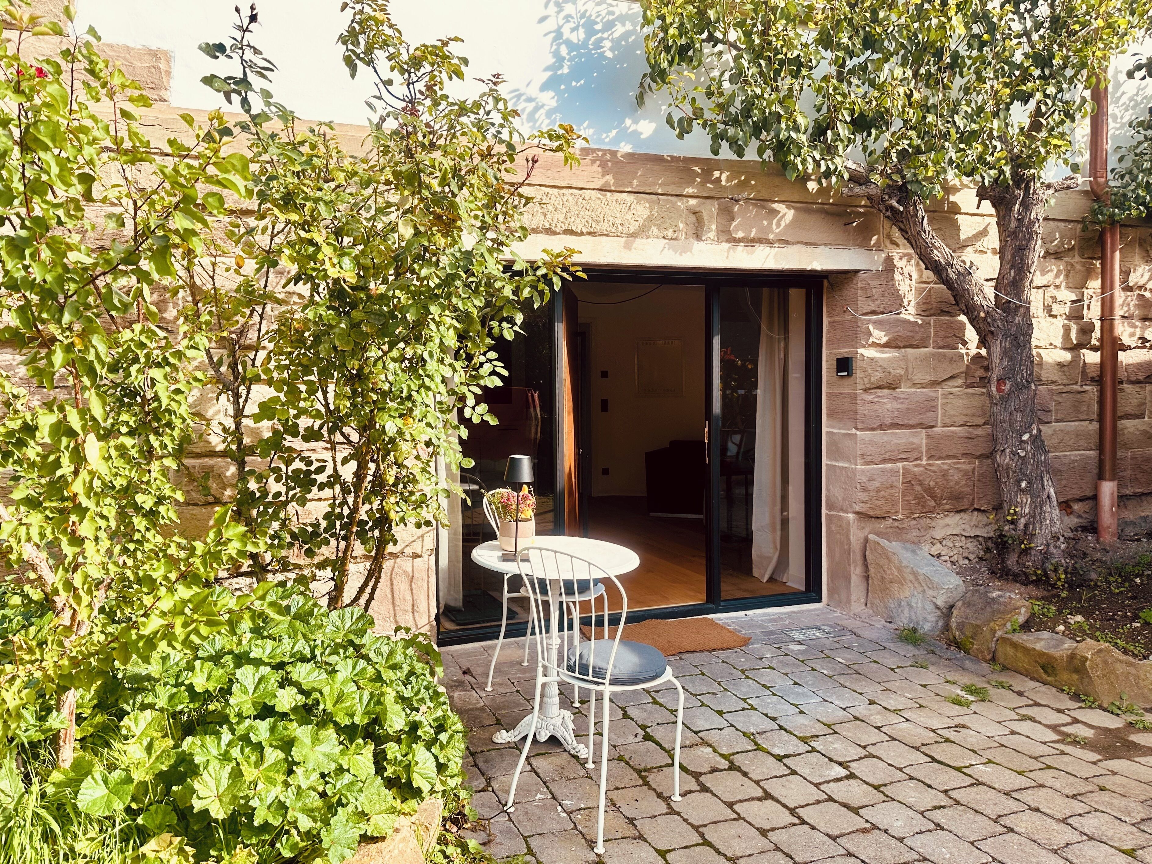 Un patio con una mesa y sillas, una puerta que da al interior y un muro de piedra.