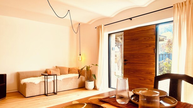 Una sala de estar moderna con un sofá, una mesa de comedor dispuesta con platos y un florero, una lámpara colgante y vistas a través de un ventanal.