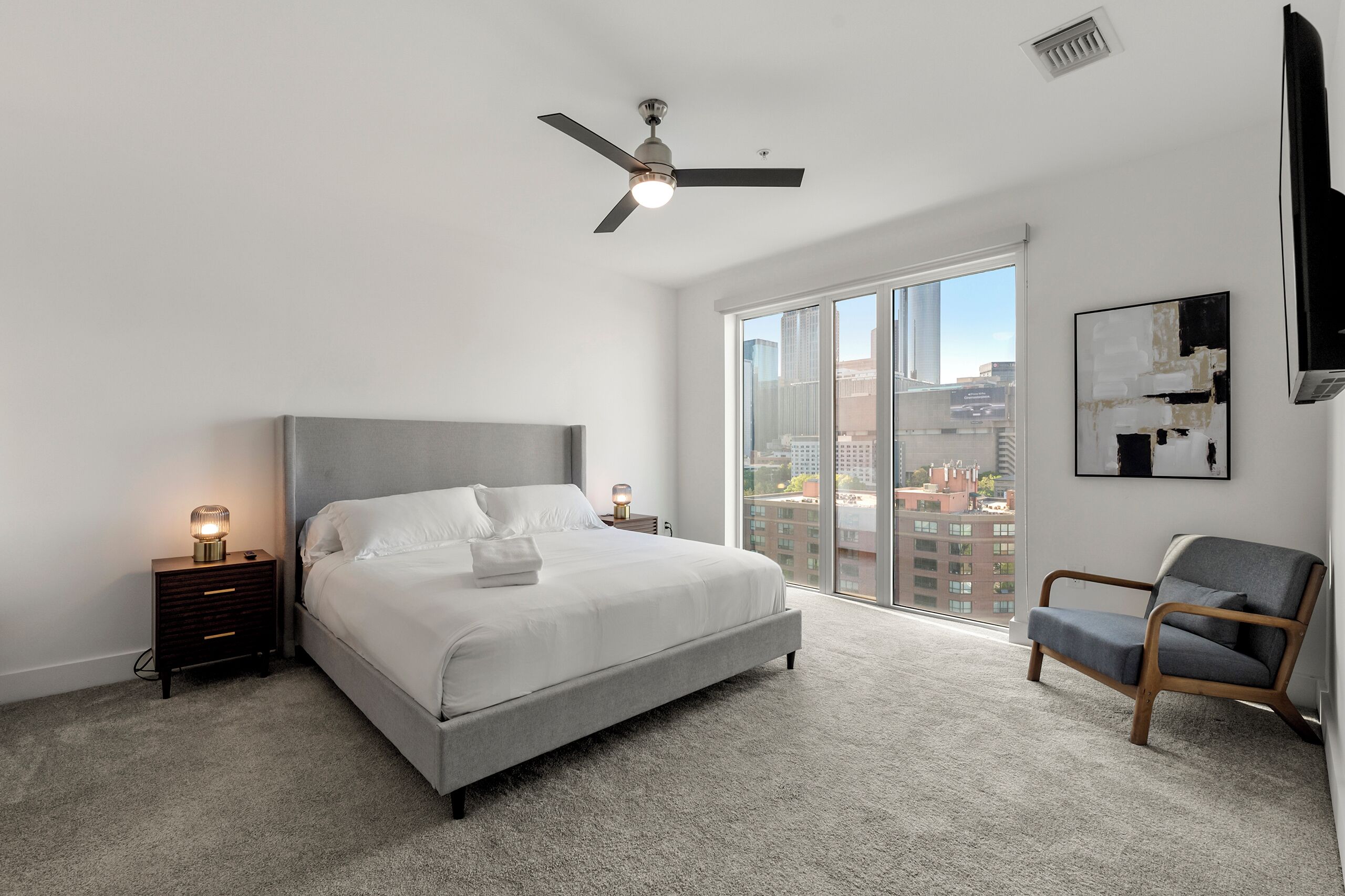 A modern bedroom with a large bed, a ceiling fan, a nightstand with a lamp, a chair, and a city view through the window.