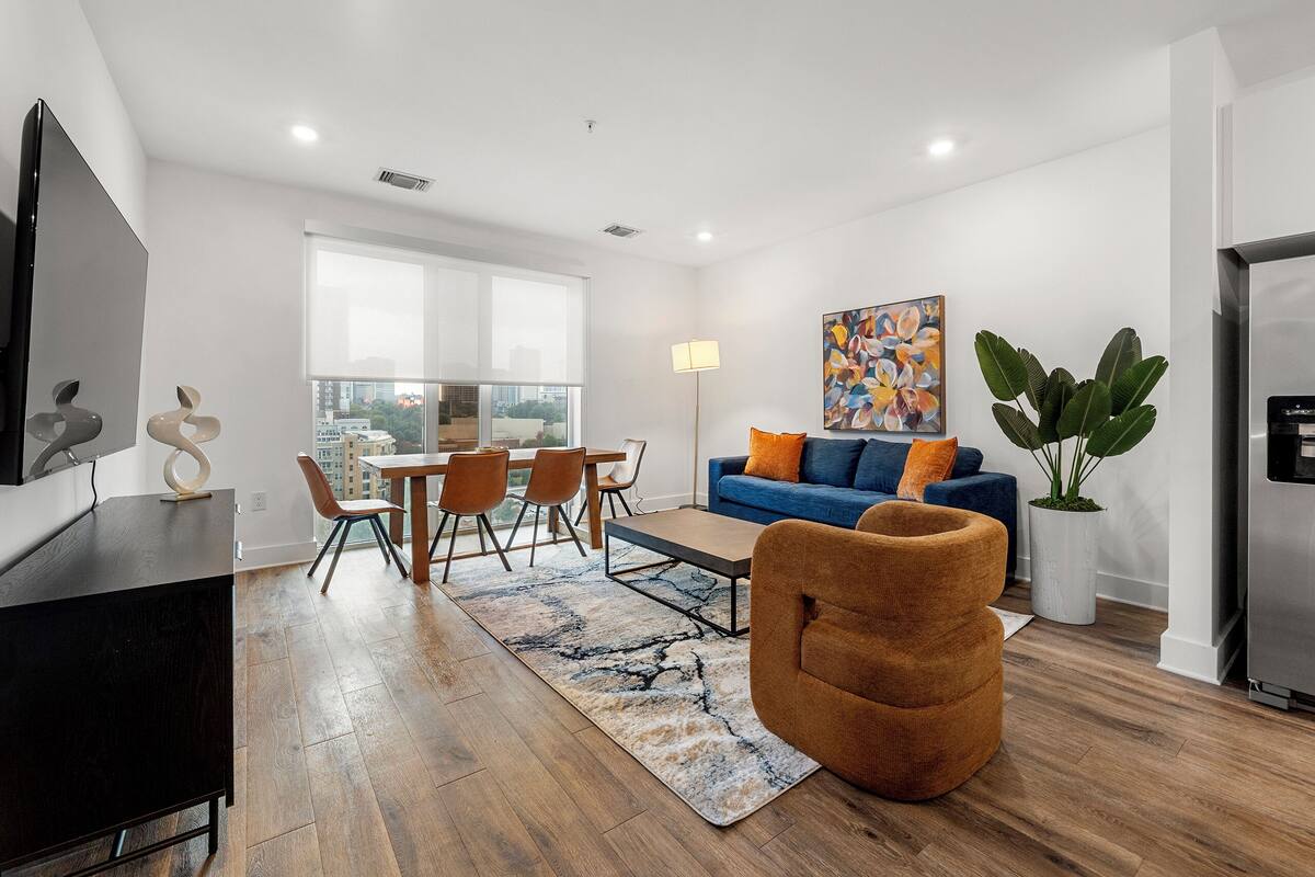 A modern living room with a dining area, a blue sofa, and a large window offering a city view.