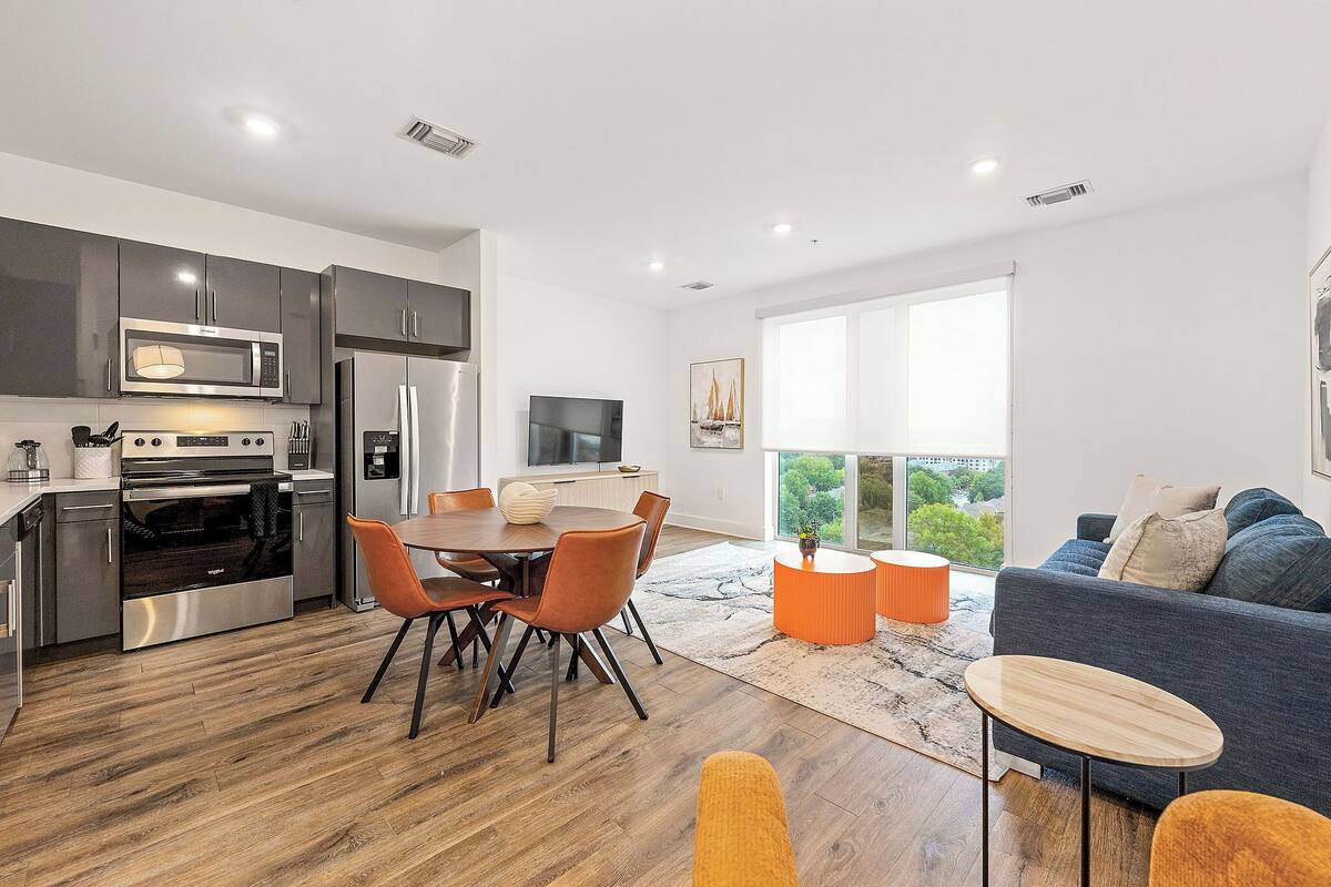 A modern kitchen with stainless steel appliances, a dining area with wooden flooring, and a living room with a sofa and a large window.