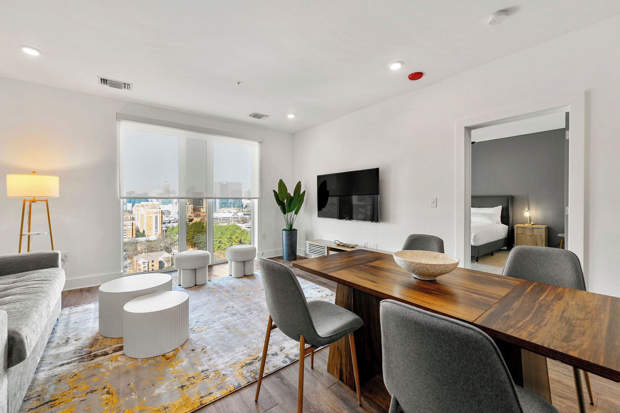 A modern living room with a wooden dining table, grey upholstered chairs, a large window with a city view, a flat-screen TV mounted on the wall, and a potted plant.