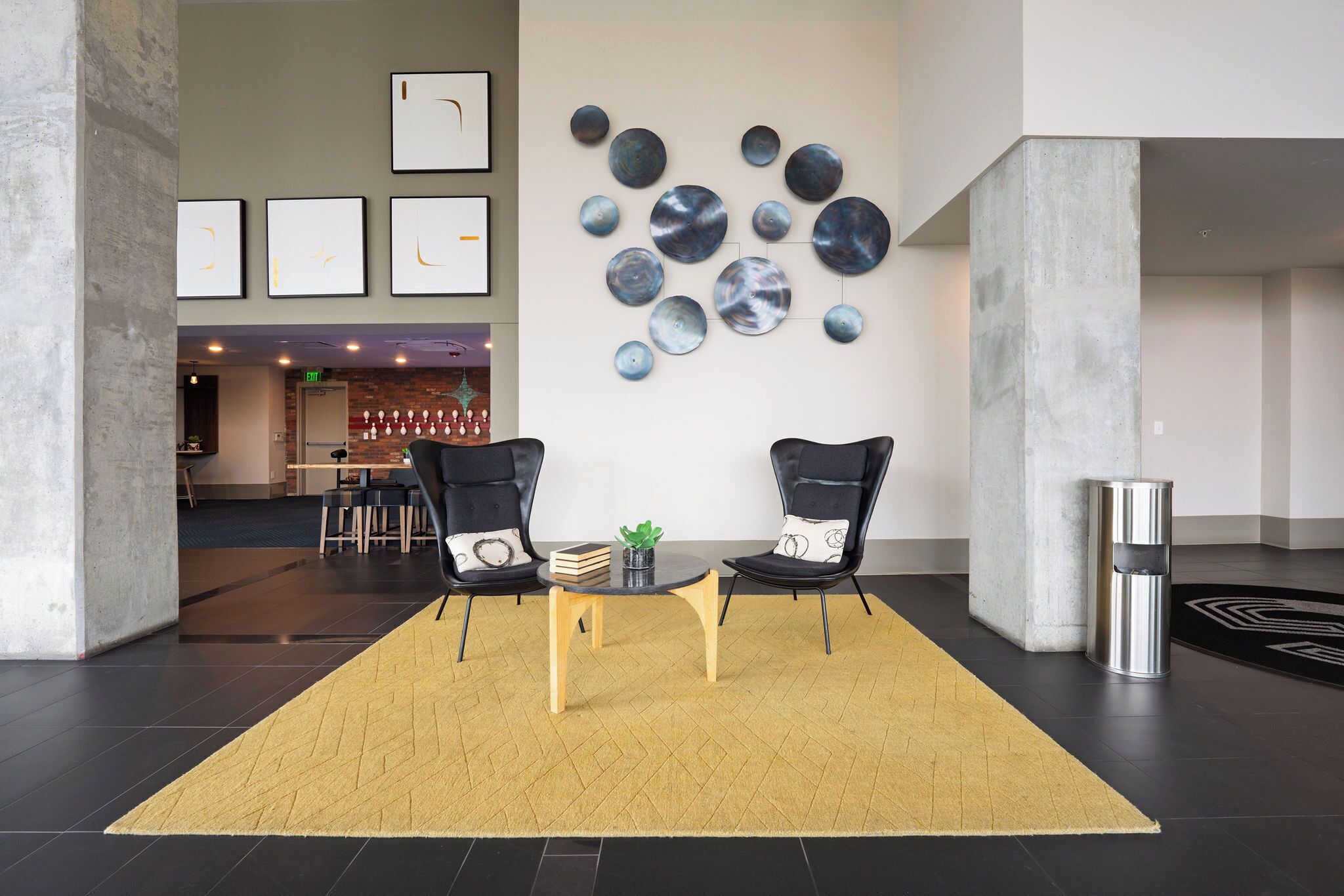 A lobby with two black chairs, a small table with a plant, and a yellow rug.