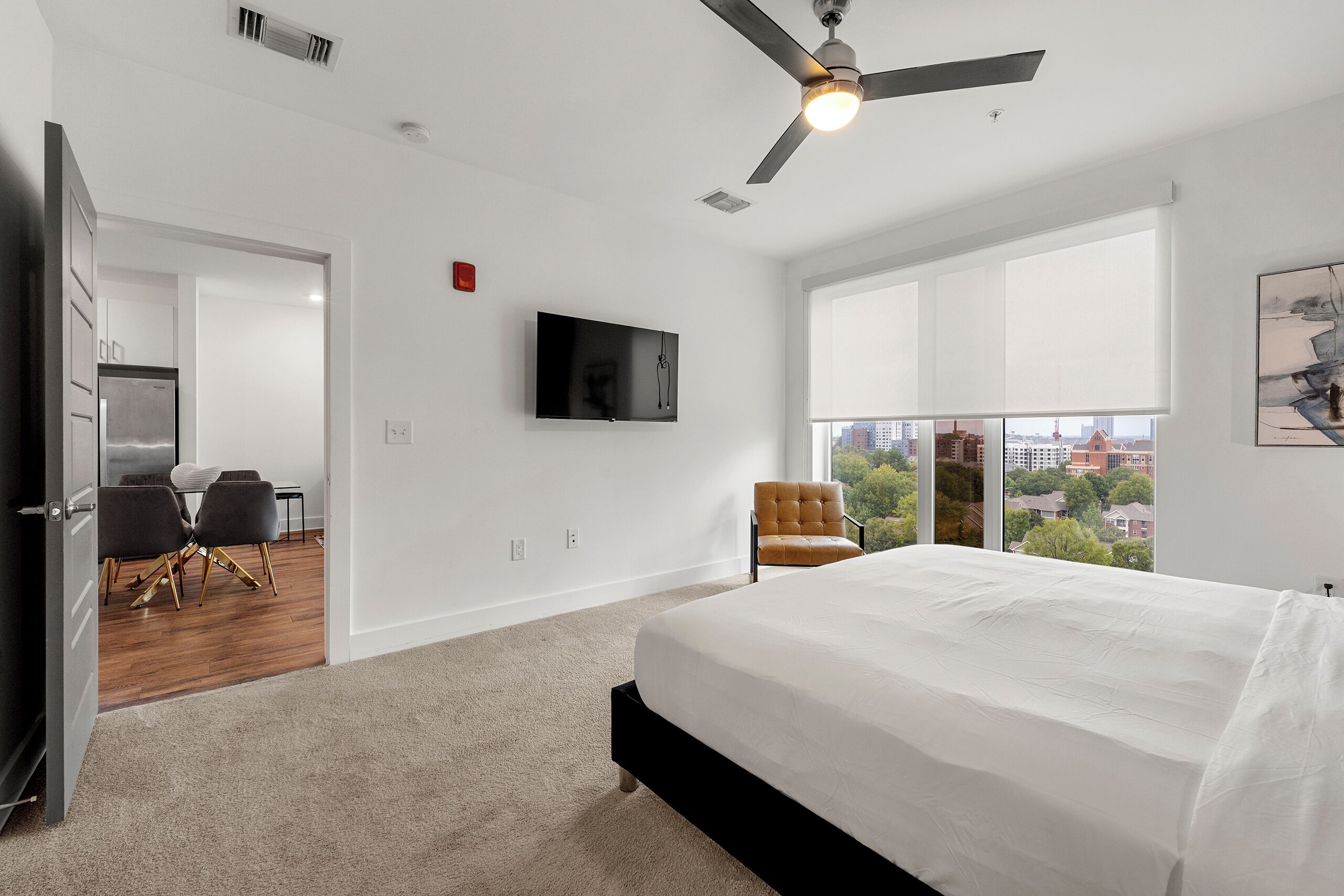 A spacious bedroom with a large bed, a ceiling fan, a flat-screen TV, and a city view through large windows.