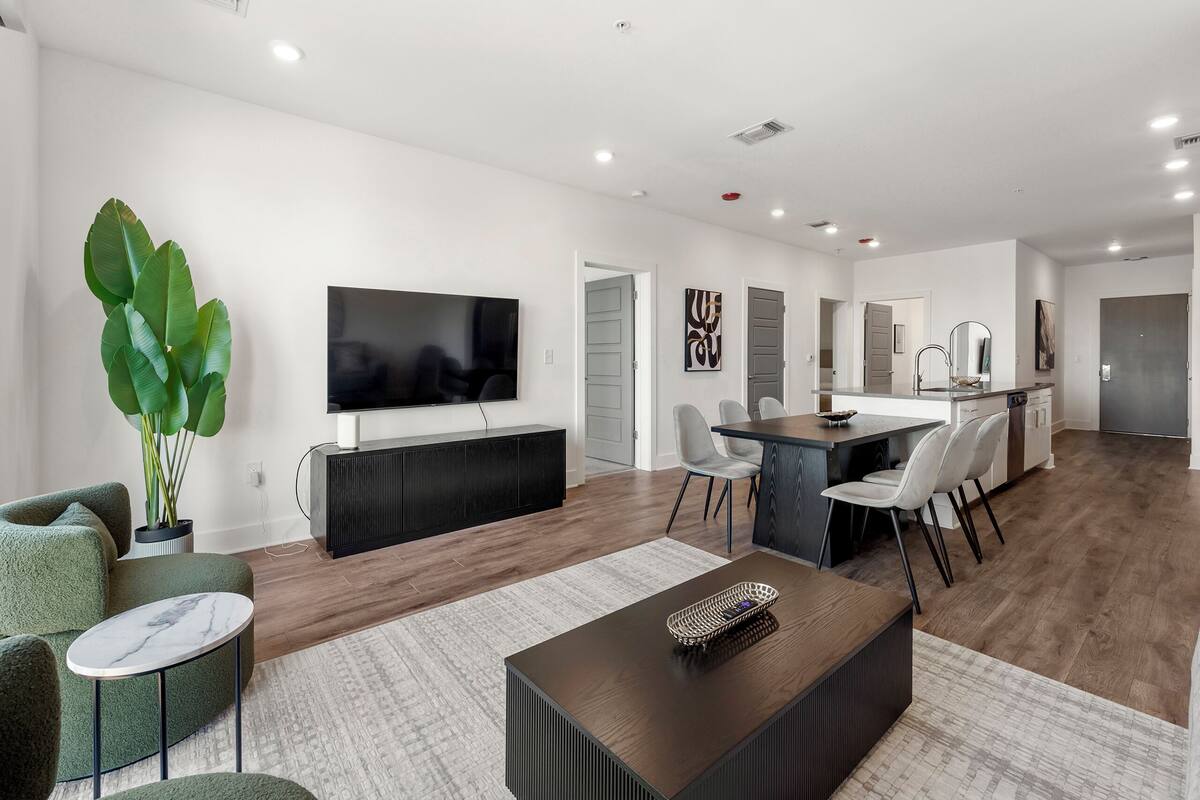 A modern living room with a flat-screen TV, green sofa, and a dining area with a kitchen in the background.