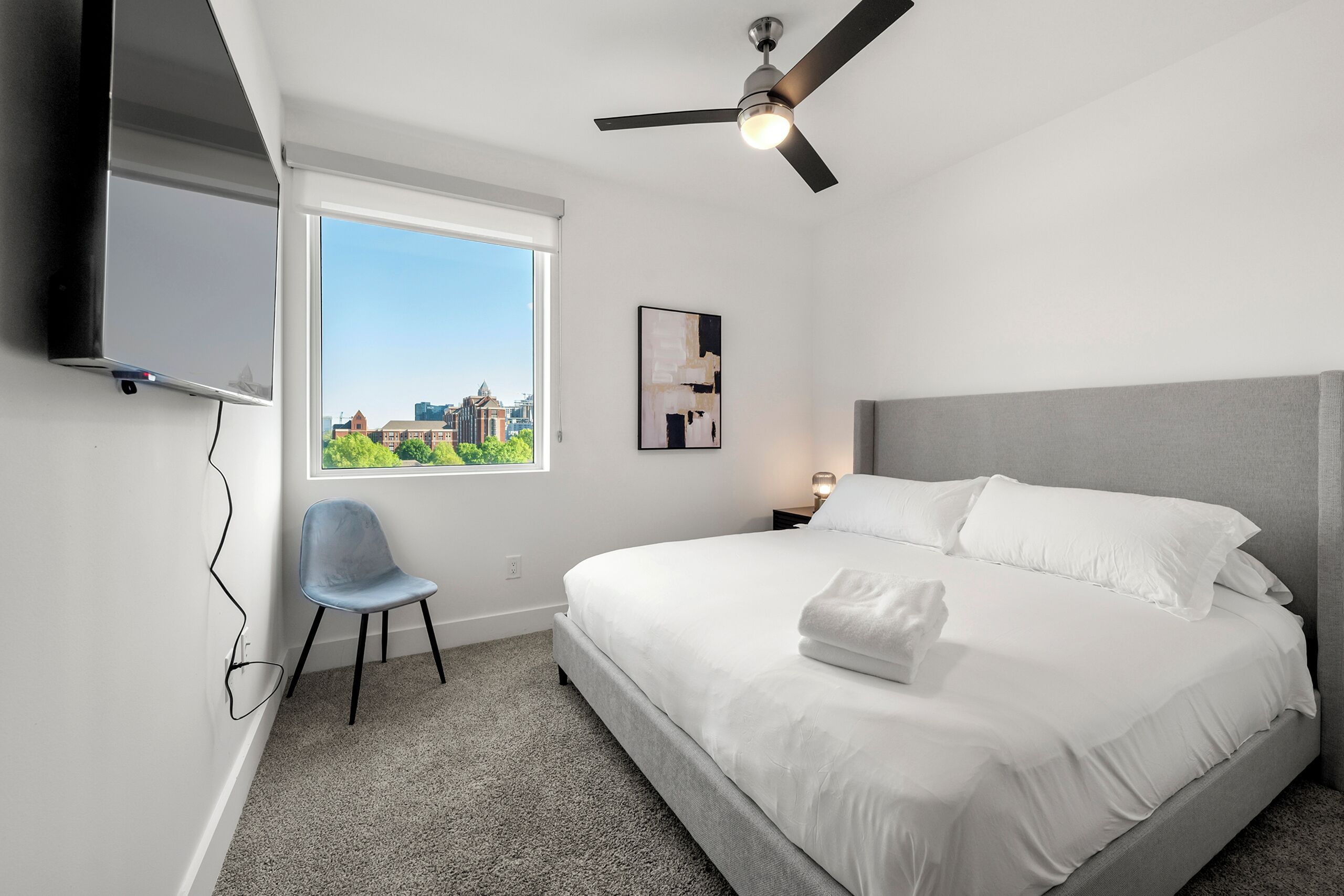 Une chambre d’hôtel moderne dotée d’un grand lit, d’un ventilateur de plafond, d’un téléviseur fixé au mur, d’une chaise et d’une fenêtre donnant sur la ville.