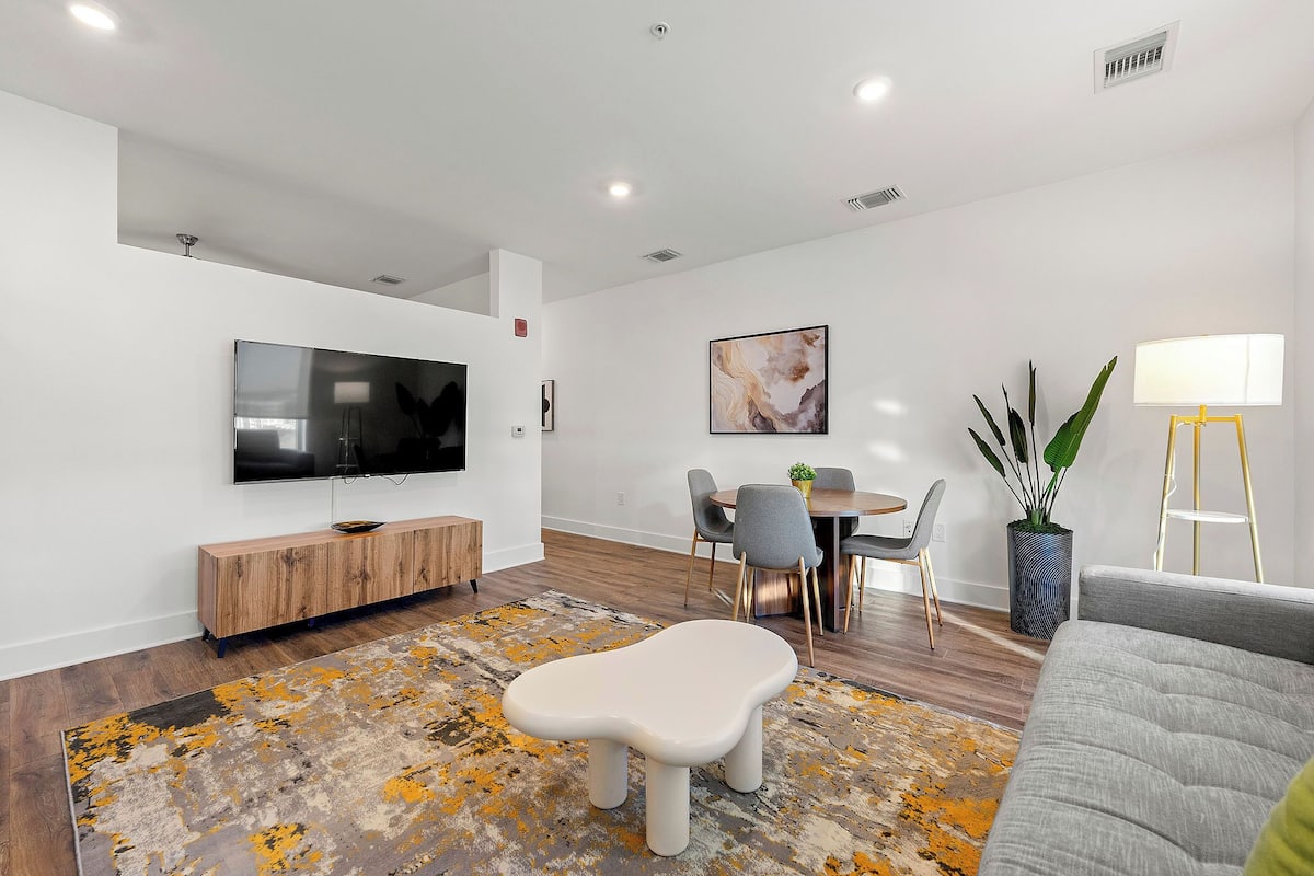 A modern living room with a flat-screen TV, a wooden console, a dining area with a table and chairs, and a grey sofa.