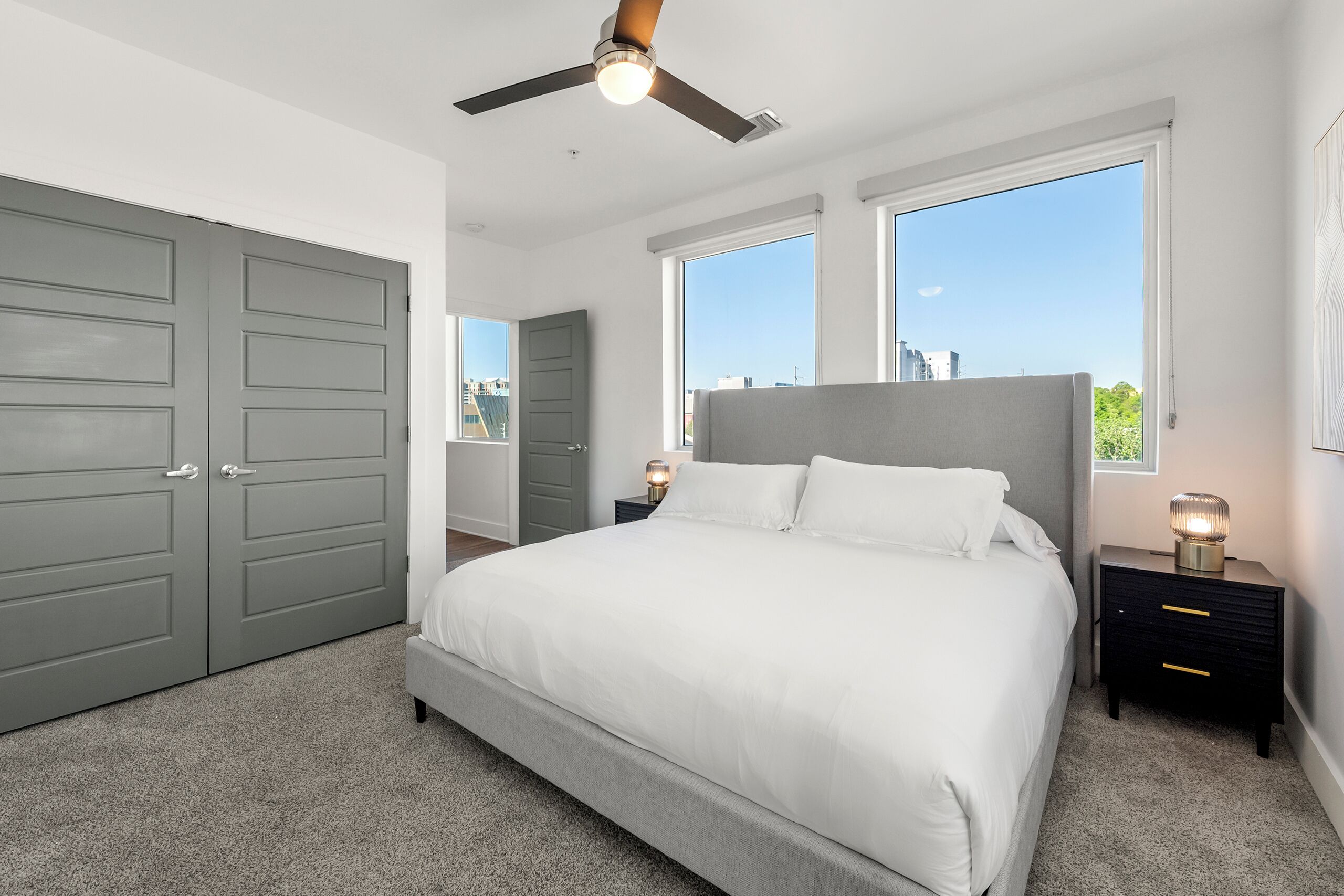 A bedroom with a bed, two bedside tables, a ceiling fan, and large windows.