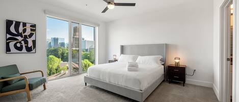 A bedroom with a large bed, a ceiling fan, a nightstand with a lamp, a green chair, and a view of the city through large windows.
